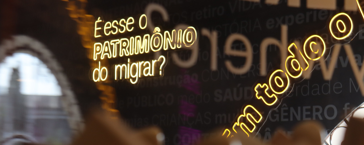 Na imagem, consta o letreiro "É esse o patrimônio do migrar?" em letras neon.